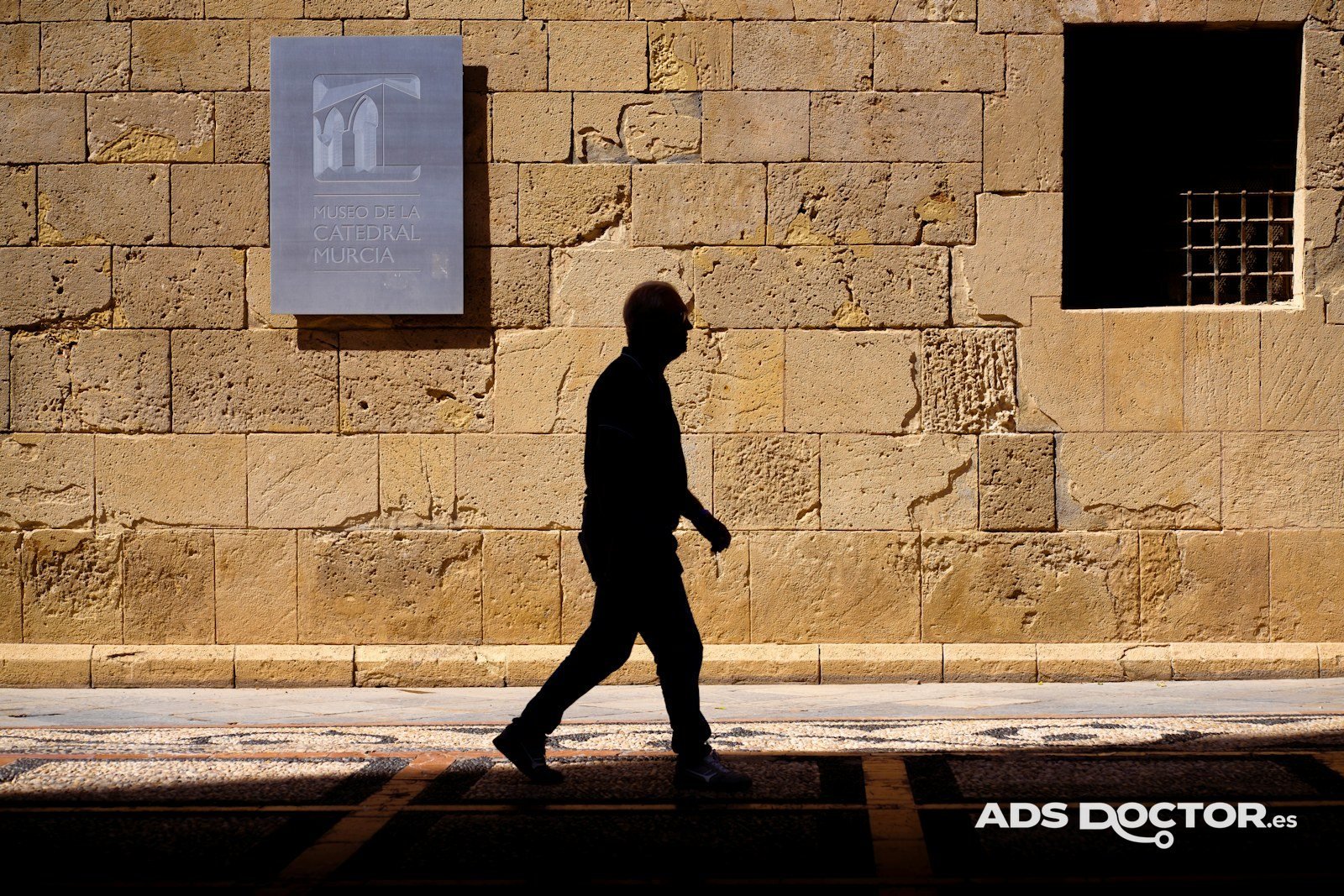 Hombre caminando en silueta junto a un muro de piedra iluminado por el sol